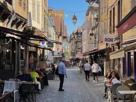 People walking on an old narrow and busy street in Troyes with blue sky and various shops and cafes lining the street