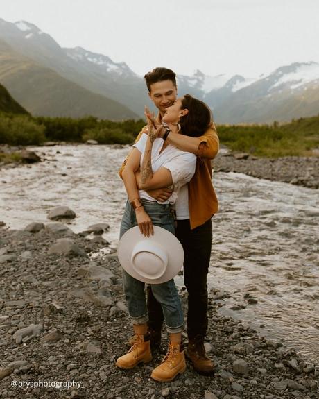 engagement photo captions love couple standing near the mountain river girl holding travel hat is her hand