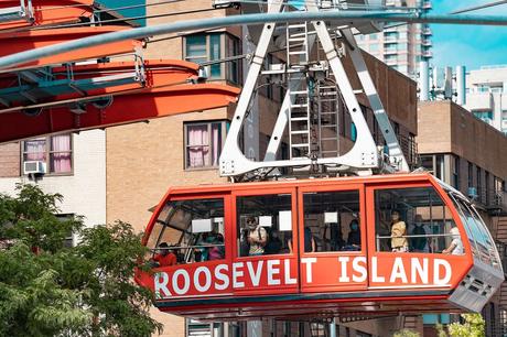 Red aerial tramway cable car with the words ROOSEVELT ISLAND painted in white containing people with buildings beyond