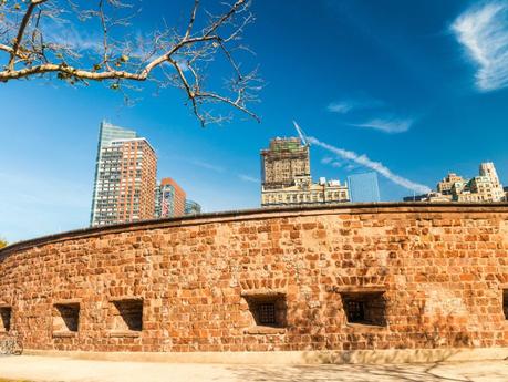 Orange stone exterior of Castle Clinton with several square windows and modern tall buildings beyond underneath a blue sky