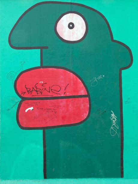 Close-up shot of graffiti on a section of the Berlin Wall in NYC showing a cartoon image of a surreal green head with red lips on a light green background