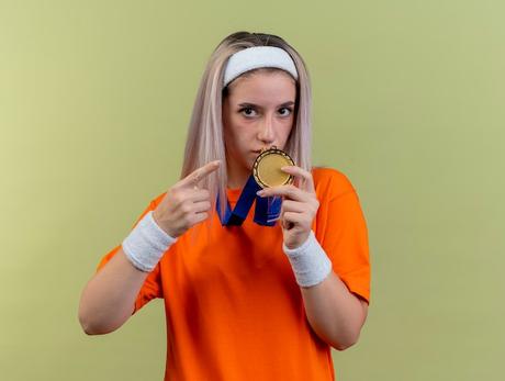 Free photo confident young caucasian sporty girl with braces wearing headband and wristbands holds and points at gold medal