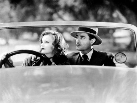 Greta Garbo and John Gilbert sitting in a convertible car.