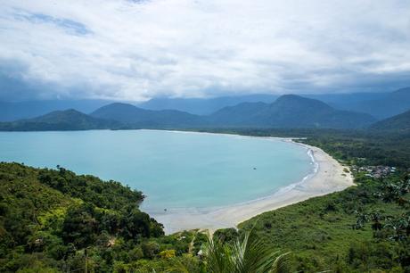 A panoramic view of a turquoise bay surrounded by forested hills and mountains, with a sandy beach stretching along the coast.