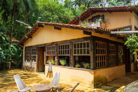 A rustic house with large windows, surrounded by lush greenery and tropical plants, showcasing traditional Brazilian architecture.