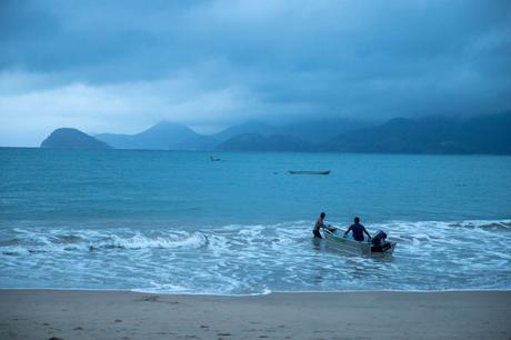 Two people pushing a small motorboat into the sea from the shore, with distant mountains and a cloudy sky in the background.