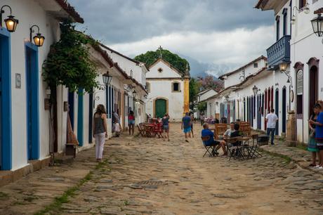 A cobblestone street in the quaint old town of Paraty with white colonial buildings, colorful doors, and people walking or sitting at outdoor cafes.