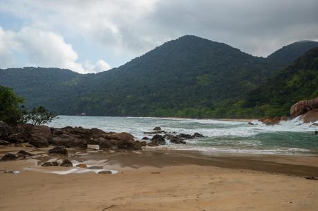 A quiet beach with rocky outcrops and waves gently crashing against the shore, set against a backdrop of forested hills.