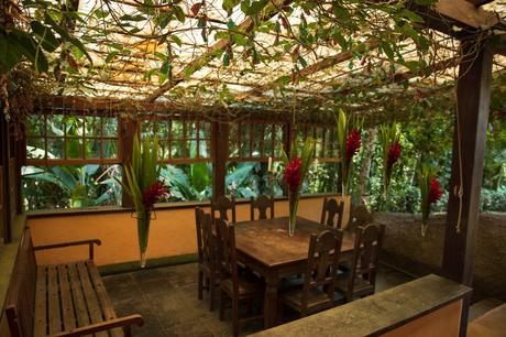 A covered outdoor dining area adorned with hanging red flowers and wooden furniture, framed by leafy green plants.