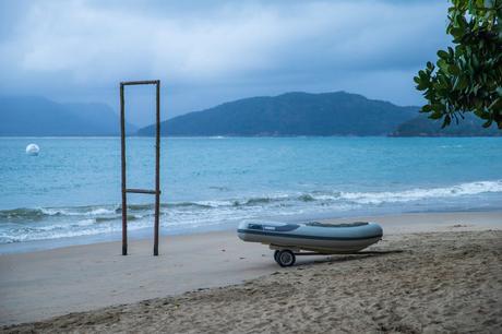 A small inflatable boat on a sandy beach with calm turquoise water in the background, framed by a wooden structure and leafy branches under an overcast sky.