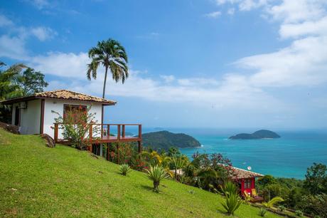 A small house with a red roof on a grassy hillside, surrounded by tropical plants and a single tall palm tree, with an ocean view in the background.