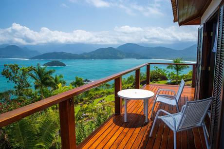 A wooden deck with two chairs and a round table overlooking a scenic landscape of green foliage, the ocean, and distant mountains under a bright, clear sky.