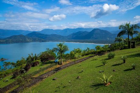 A scenic view of a lush green hillside with tropical plants and a pathway leading down toward a blue ocean bay surrounded by forested mountains under a partly cloudy sky.