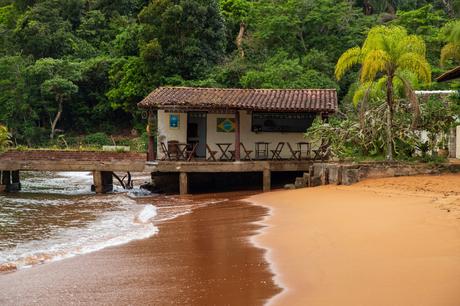 A small beachside bar with a tiled roof on stilts above the water, surrounded by lush greenery, and the sand blending into the waves.