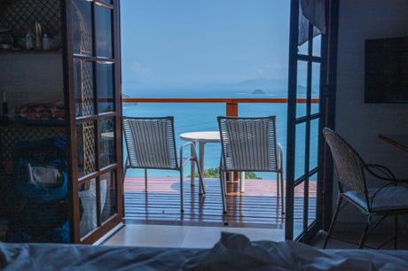 A cozy balcony view from a room, featuring two white wicker chairs and a small table overlooking a blue ocean with distant islands under a bright sky.