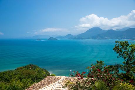 A panoramic view of an expansive ocean, surrounded by multiple small green islands, with vibrant tropical vegetation in the foreground and distant mountains under a blue sky.