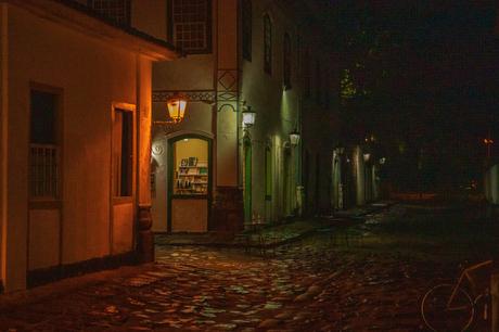 An illuminated narrow street in the Brazilian town of Paraty at night, with warm streetlights casting a glow on the wet cobblestone pavement and the facades of historic buildings.