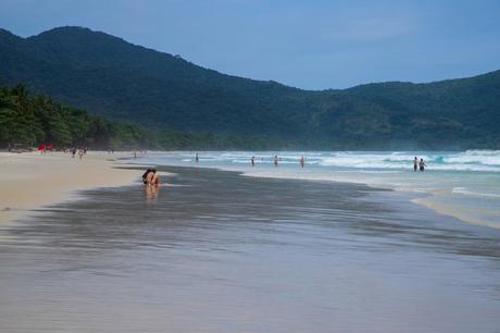 A wide, sandy beach on Ilha Grande with waves and people enjoying the water, bordered by green forested hills under a bright blue sky.