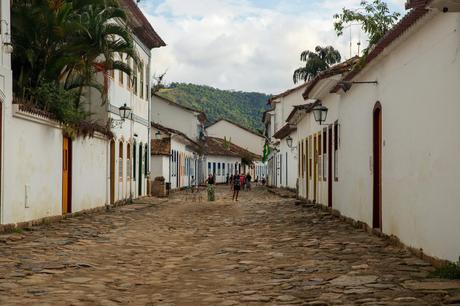 A quiet, cobblestone street in the historic Brazilian town of Paraty, lined with white colonial buildings and mountain views in the background.