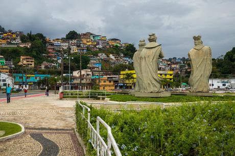 A large public square with three monumental statues facing a hillside covered in colorful houses under a cloudy sky.