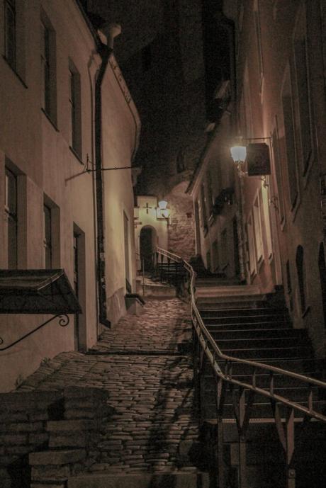 A similar photo, with slightly different lighting effects, of the same narrow, cobblestone alleyway in the old town of Tallinn, lit by dim street lamps. The alley has a gentle upward slope, bordered by old buildings with wooden shutters.