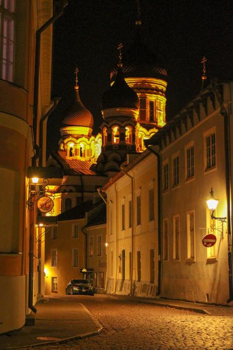 A nighttime view of an old street in Tallinn with ornate church domes lit up in warm yellow light. The street is lined with traditional buildings and vintage street lamps.