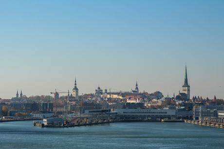 A panoramic view of Tallinn, Estonia, from the sea, showcasing the city's skyline with its medieval towers and spires. The harbor is in the foreground with cranes, docks, and buildings, while the historic city center rises in the background against a clear blue sky.