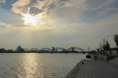 A serene view of the Daugava River at sunset, with a silhouetted bridge and modern cityscape under a dramatic sky with golden hues and scattered clouds.