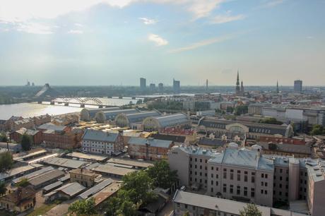 An aerial view of Riga, Latvia, showcasing the city's skyline with church spires, bridges, and modern buildings along the Daugava River under a partly cloudy sky.