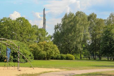 A serene park with lush greenery, trees, and an open grassy area. In the background, the tall, slender Victory Monument rises above the treetops, with a golden star at its peak.