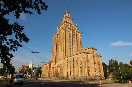 A tall Stalinist-era skyscraper in Riga, known as the Latvian Academy of Sciences, standing against a bright blue sky, framed by leafy trees in the foreground.