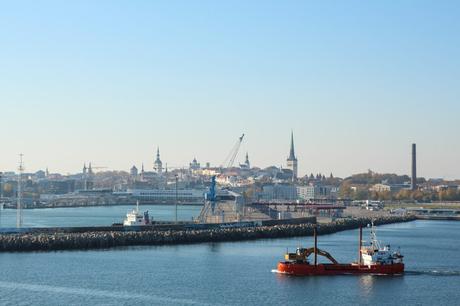 A view of Tallinn Harbour, featuring a ship navigating the calm waters. The city's skyline, dotted with medieval church spires and modern buildings, is visible in the background, under a bright, cloudless sky.