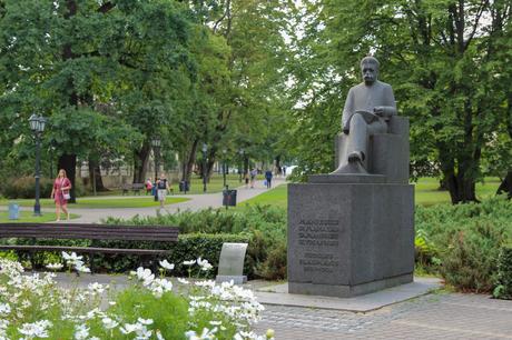 A prominent stone statue of a seated figure, set in a well-maintained park with pathways, benches, and a few pedestrians enjoying the surroundings. Trees and greenery frame the scene.