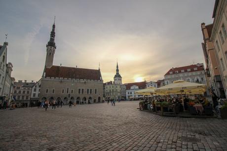 A cobblestone square in a historic town, with people walking and dining outdoors under large umbrellas. The square is framed by a tall church tower and other buildings, with the sun setting in the background.