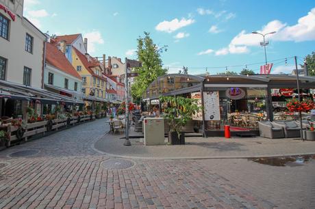 A picturesque cobblestone street lined with colorful cafes and restaurants, outdoor seating, and flower arrangements under a clear blue sky.