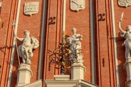 A close-up view of three white statues against the red brick facade of the House of the Blackheads in Riga. The statues depict classical figures, including Neptune with a trident and a woman holding a scroll, highlighted by gold embellishments.