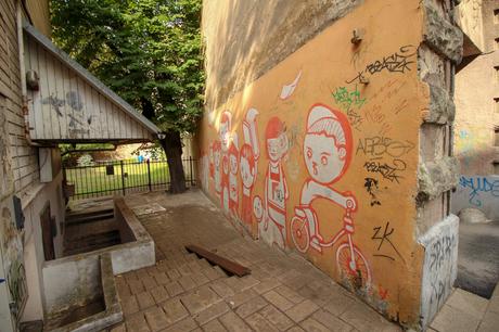 A colorful graffiti mural featuring cartoon-like characters on a peach-colored wall in a narrow alley with brick paving.