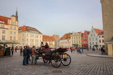 A lively square in Tallinn, Estonia, with colorful historic buildings and a horse-drawn carriage. People are gathered around, enjoying the ambiance, while the pastel-colored facades and cobblestone streets create a picturesque scene.