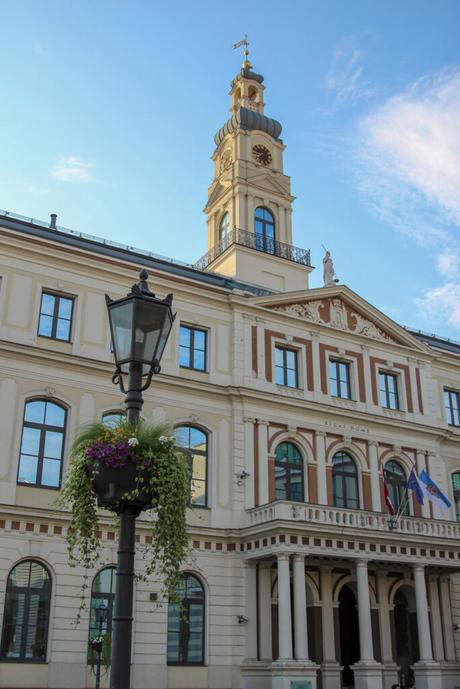 The Riga Town Hall, featuring a classical architectural style with white walls, large windows, and a tall clock tower with a dome. A street lamp adorned with flowers is in the foreground, adding to the charming cityscape.