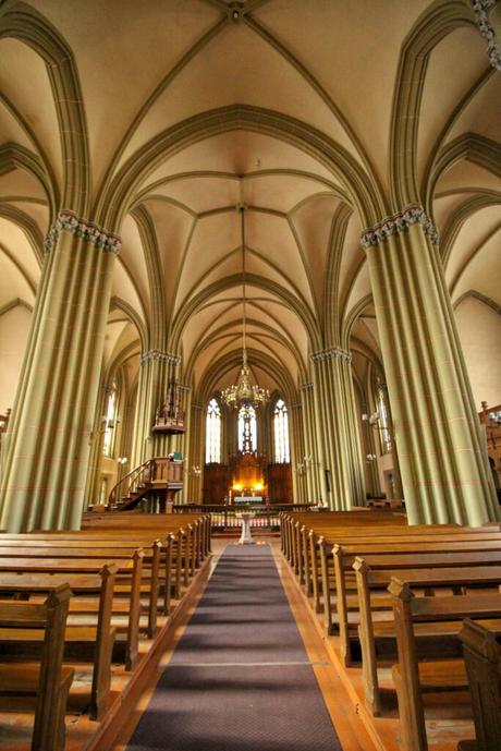 The interior of a Gothic-style church with high, arched ceilings, tall columns, and wooden pews leading toward the altar illuminated by soft light.