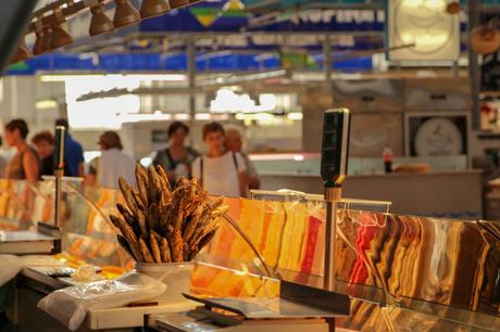 A close-up of a market stall displaying a large bunch of dried fish in a bowl, with a background of blurred customers and warm lighting reflecting off glass surfaces.