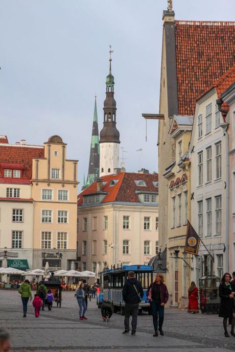 A bustling street scene in the Baltics with pastel-colored buildings, a tram, and people walking. A tall church spire rises in the background under a cloudy sky.