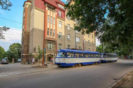 A blue tram is parked near a large, ornate building with a red and beige exterior, while pedestrians walk along the street.