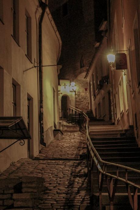 A narrow, cobblestone alleyway in the old town of Tallinn, lit by dim street lamps. The alley has a gentle upward slope, bordered by old buildings with wooden shutters.