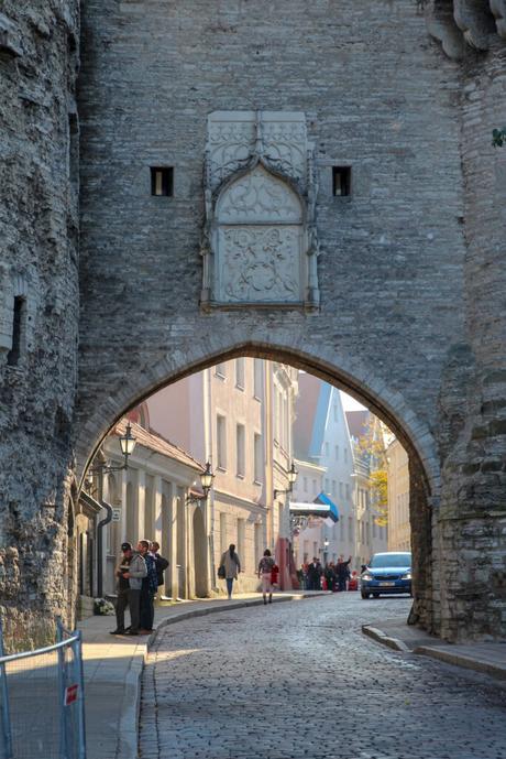 An arched stone gateway in the old town of Tallinn, Estonia, with a narrow cobblestone street leading through it. People are gathered near the entrance, and a car is parked nearby.