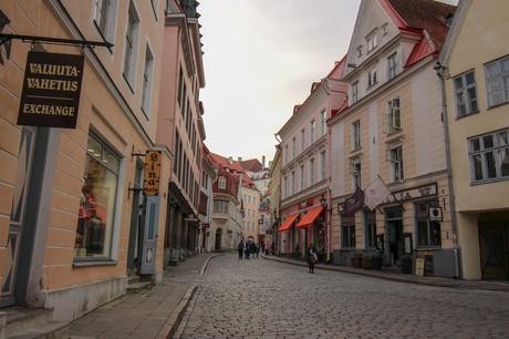 A quaint, narrow street in Tallinn's Old Town, lined with pastel-colored buildings with signs for shops and cafes. The cobblestone pathway winds gently, and a few people are walking, creating a cozy and inviting atmosphere.