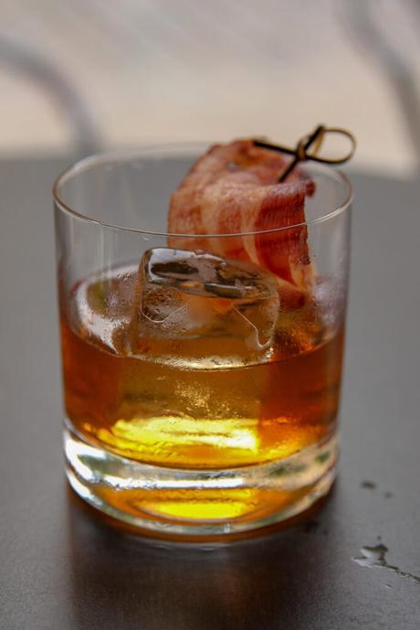 A glass of whiskey with a large ice cube, garnished with a crispy strip of bacon skewered on a toothpick.