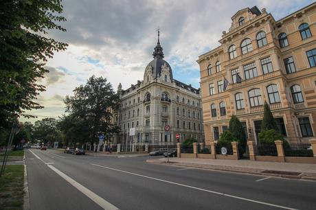 A grand, historic building with intricate architectural details on a quiet street, with cars and a few pedestrians in the background.