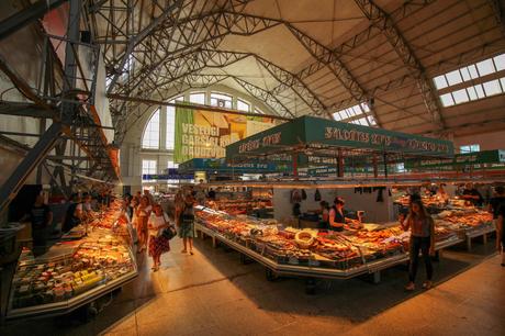 A bustling indoor market hall filled with food stalls and vendors. A high, arched ceiling with metal beams covers the expansive space, while customers browse the various offerings under bright, welcoming lights.