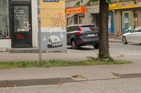 A small piece of street art depicting a stylized skull, painted on the corner of a building in an urban environment with parked cars and nearby shops.